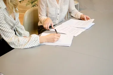 Dwie osoby przy stole podpisują i przeglądają dokumenty.