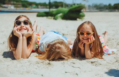 Trzy dziewczynki leżą na plaży w okularach przeciwsłonecznych i uśmiechają się.