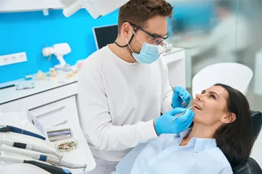 Dentysta w masce i rękawiczkach bada zęby pacjentki na fotelu.