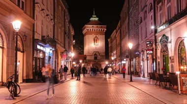 Nocna ulica z przechodniami, oświetlonymi kamienicami i zabytkową bramą na końcu.