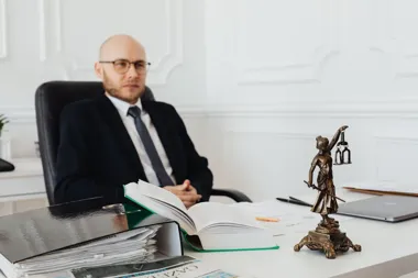 Adwokat w garniturze siedzi przy biurku z aktami, otwartą księgą i figurką Temidy.