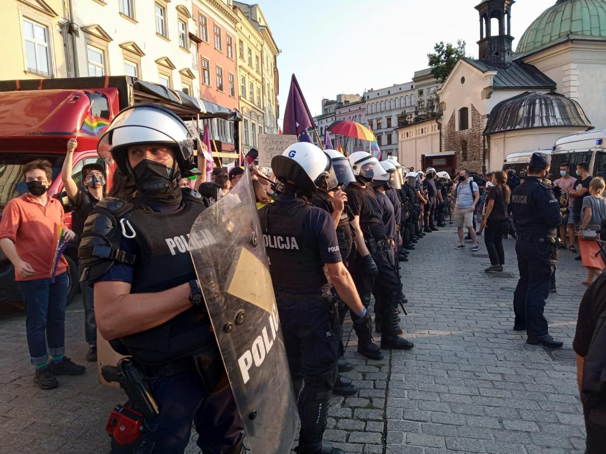 kordon kilkudziesięciu policjantów na krakowskim rynku zabezpieczający zgromadzenie publiczne. W tle osoby biorące udział w zgromadzeniuz kolorowymi emblematami oraz czerwony bus