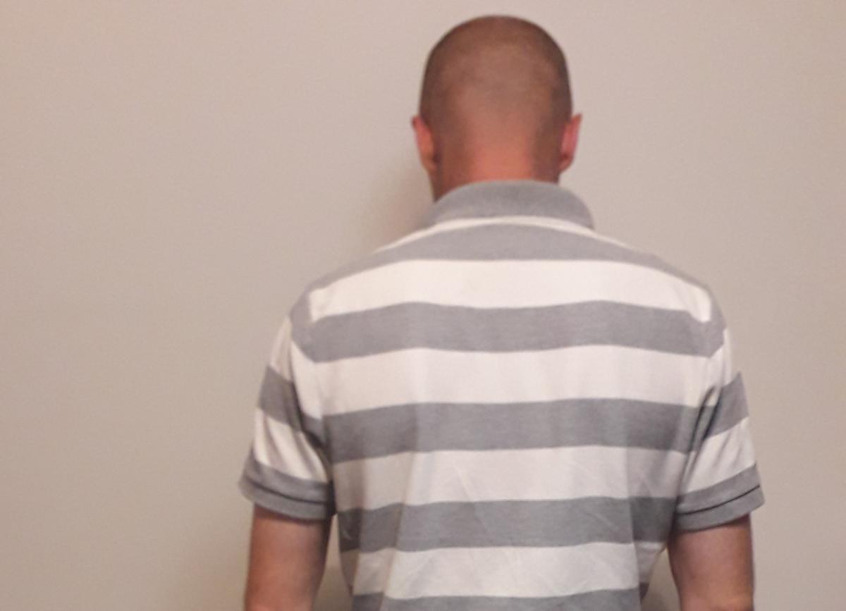 zatrzymany sprawca pobicia ubrany w białą koszulke w szare psaki stojący tyłem do zdjęcia