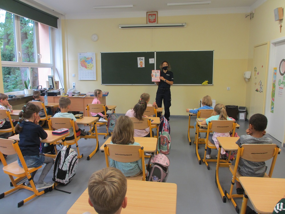 Policjantka trzymająca w ręce tablicę edukacyjną prowadzi zajęcia dla dzieci w sali lekcyjnej