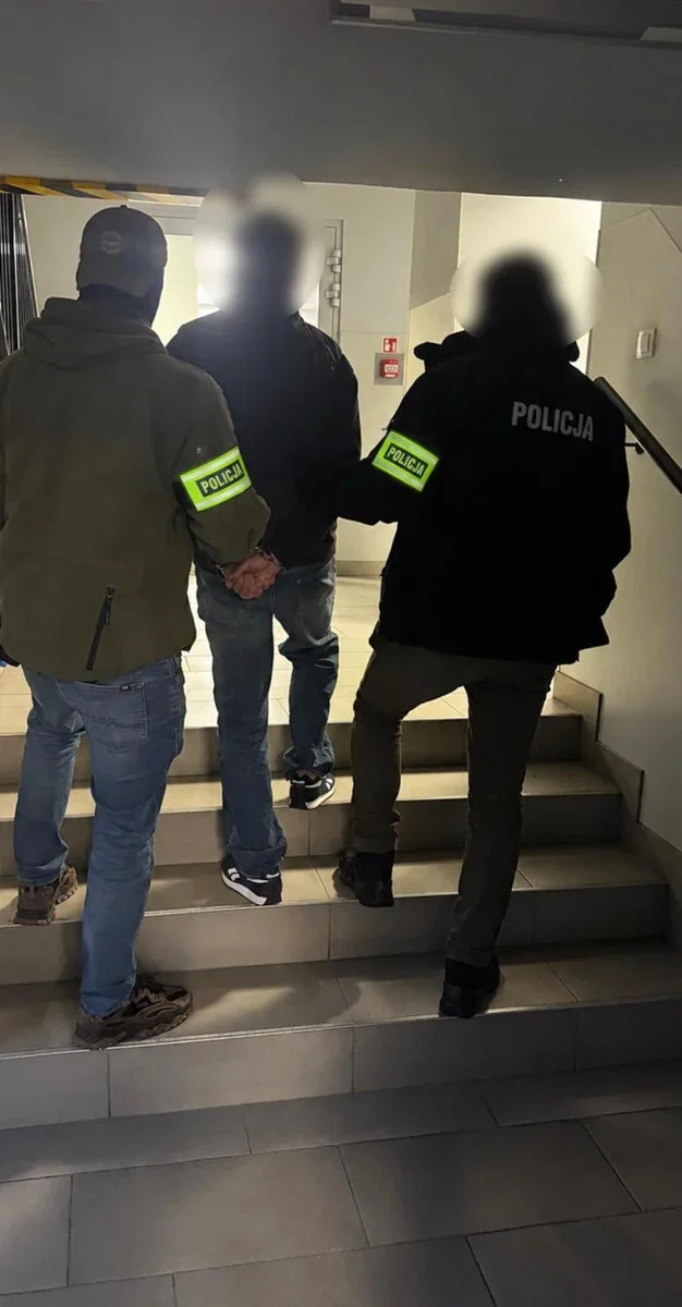 Funkcjonariusze policji prowadzą zatrzymanego mężczyznę po klatce schodowej wewnątrz budynku.