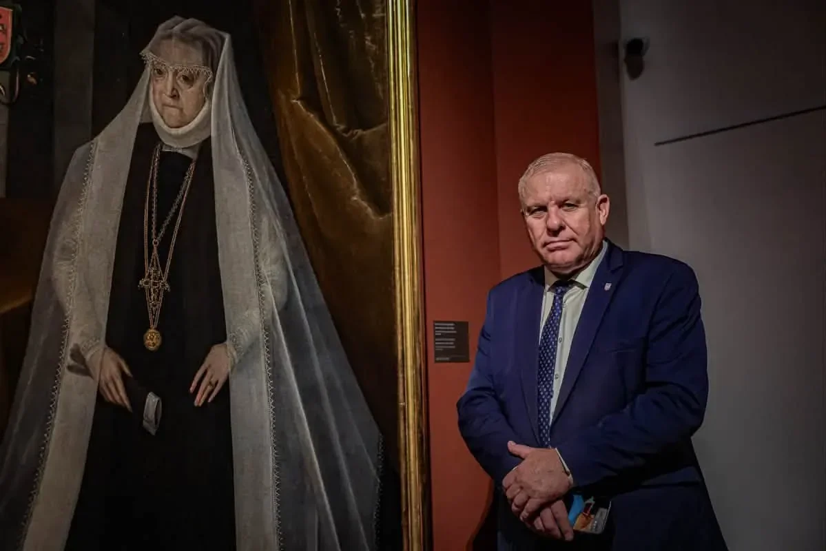 Mężczyzna w granatowym garniturze stoi obok dużego portretu kobiety w czarnej sukni i białym welonie w pomieszczeniu muzealnym.