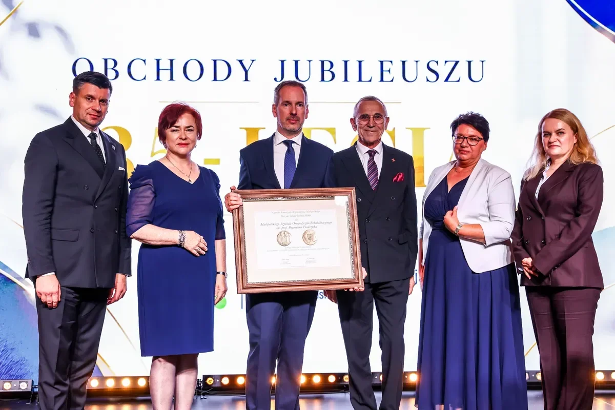 Sześć osób na scenie podczas uroczystości jubileuszu, dwóch mężczyzn wręcza lub odbiera dużą ramkę z dyplomem.