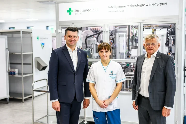 Trzy osoby pozują do zdjęcia w pomieszczeniu produkcyjnym firmy Intelimedical Poland z inteligentnymi technologiami medycznymi w tle.