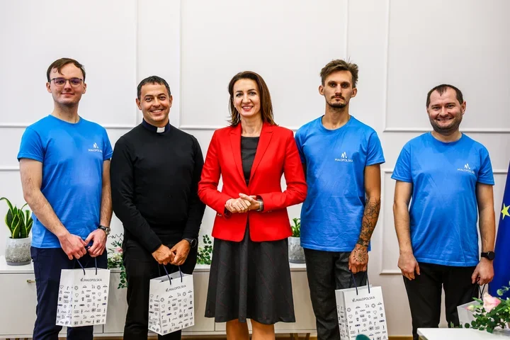 Grupa pięciu osób, w tym kobieta w czerwonej marynarce i mężczyzna w czarnym duchownym ubraniu, pozuje do zdjęcia w eleganckim pomieszczeniu; trzej mężczyźni w