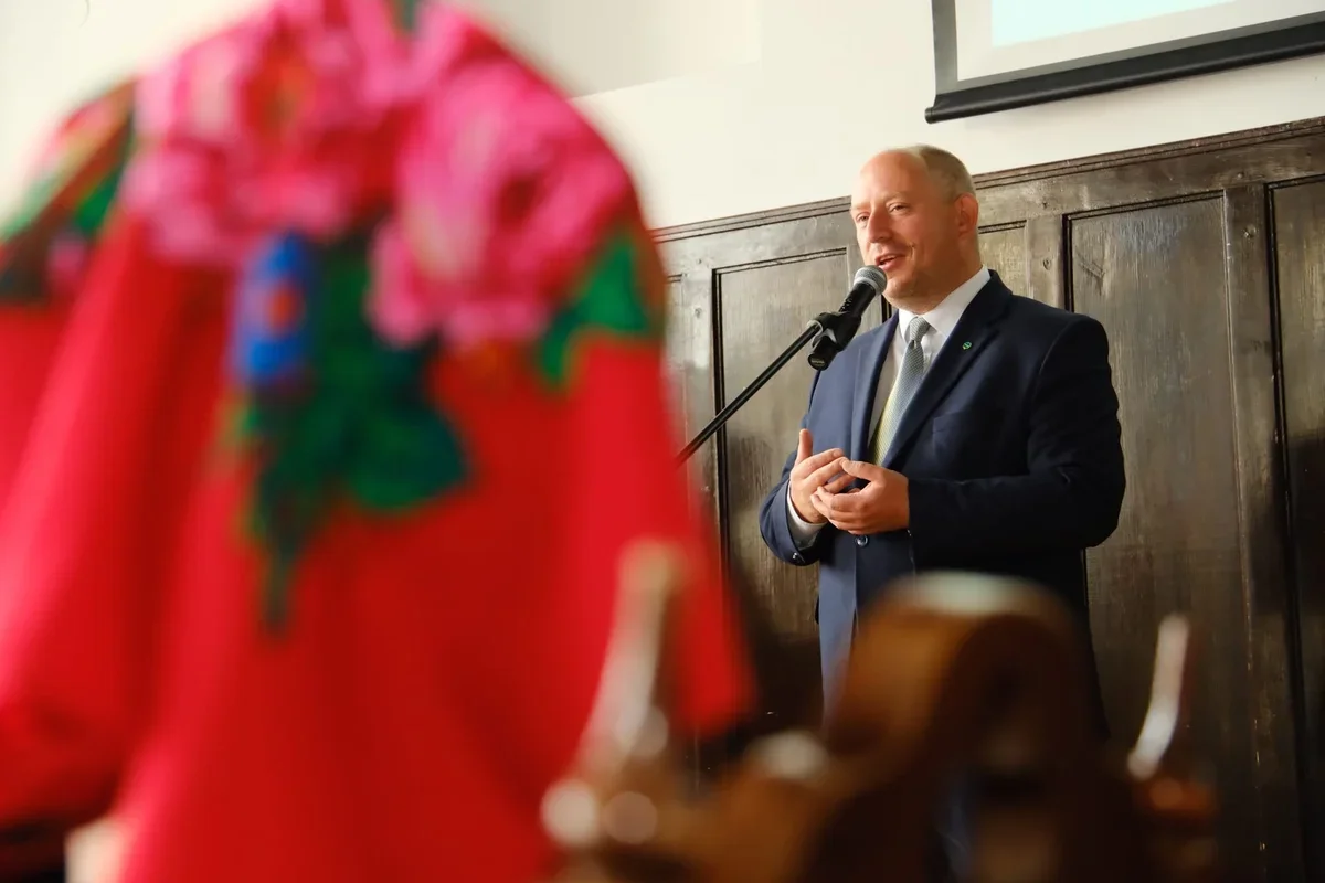 Mężczyzna w garniturze przemawia do mikrofonu podczas uroczystości w pomieszczeniu z drewnianymi panelami.