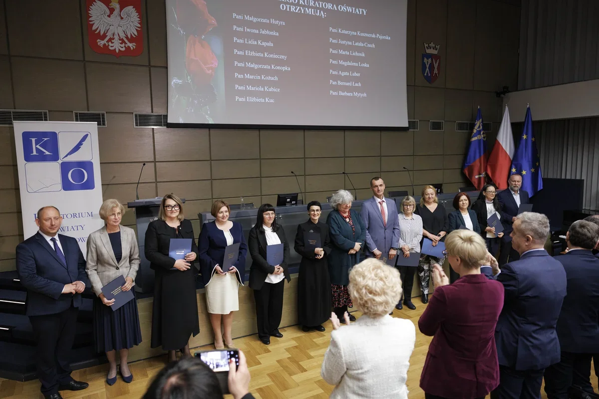 Uroczystość wręczenia nagród kuratora oświaty w sali konferencyjnej, podczas której wyróżnione osoby stoją na scenie z dyplomami, a publiczność im aplauduje.