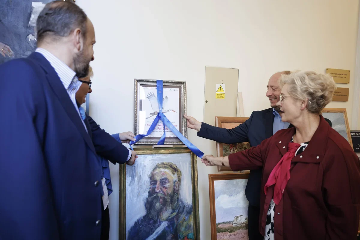 Cztery osoby otwierają niebieską wstęgą uroczyście oprawiony plakat z bocianem, wiszący nad portretem mężczyzny w pomieszczeniu z obrazami.
