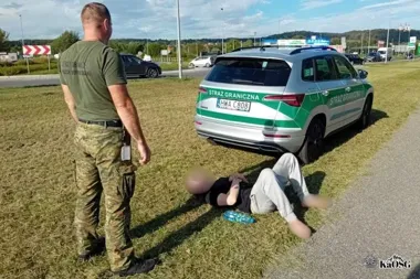 Funkcjonariusz Straży Granicznej stoi na trawie przy radiowozie obok leżącej na ziemi osoby, na drodze w tle widoczny ruch pojazdów.
