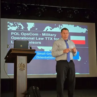 Mężczyzna w koszuli i krawacie przemawia przy pulpicie podczas prezentacji z napisem „POL OpsCom - Military Operational Law TTX for Legal Advisors”.