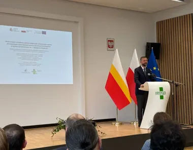 Mężczyzna w garniturze przemawia z pulpitu w sali konferencyjnej obok flag polskiej, małopolskiej i unijnej, z projekcją informacji o inwestycji w Szpitalu Rydy
