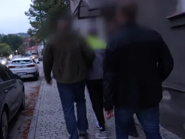 Trzech mężczyzn spaceruje brukowanym chodnikiem wzdłuż ulicy w mieście o zmierzchu, po lewej stronie zaparkowane samochody.