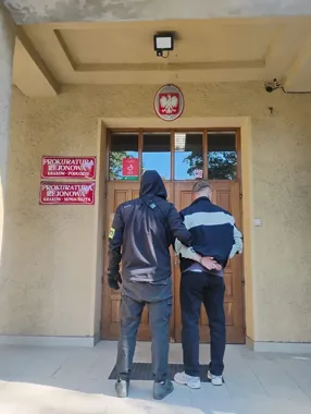 Funkcjonariusz w kurtce z kapturem prowadzi mężczyznę w kajdankach do drzwi Prokuratury Rejonowej Kraków-Podgórze i Kraków-Nowa Huta.