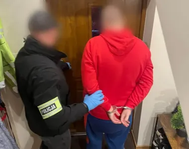 Funkcjonariusz policji w niebieskich rękawiczkach trzyma mężczyznę w czerwonej bluzie z rękoma skutymi kajdankami w przedpokoju mieszkania.