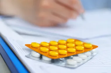 Blistry z tabletkami leżące na dokumentacji medycznej, w tle rozmyta dłoń z długopisem.