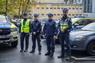 Czworo policjantów w mundurach i kamizelkach odblaskowych stoi na parkingu między policyjnymi radiowozami.