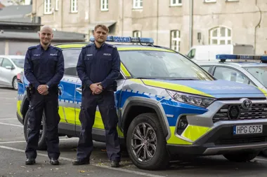 Dwóch policjantów w mundurach stoi przed policyjnym radiowozem marki Toyota zaparkowanym na ulicy.