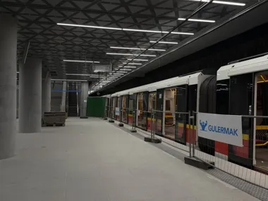 Pociąg metra stoi na nowoczesnej, częściowo wykończonej stacji podziemnej, przy peronie widoczne są betonowe kolumny, sufit z geometrycznym wzorem oraz ogrodzen