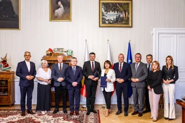 Grupa osób stoi w pomieszczeniu na tle flag Polski i Unii Europejskiej.