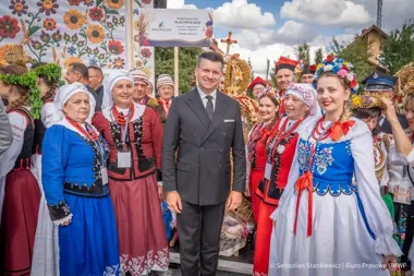 Uczestnicy w strojach ludowych pozują do zdjęcia z mężczyzną w garniturze podczas plenerowej uroczystości dożynkowej w województwie małopolskim.