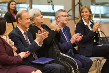 Grupa osób w formalnym stroju siedzi na krzesłach w sali konferencyjnej i klaska.