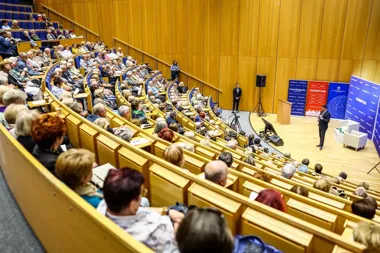 Mężczyzna w garniturze przemawia do licznej publiczności zgromadzonej w auli z drewnianymi ławkami, przy banerach medialnych i instytucjonalnych na scenie.