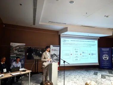 Mężczyzna w jasnym garniturze wygłasza prezentację przy pulpicie w sali konferencyjnej, za nim wyświetlona jest slajd z diagramem instytucji.