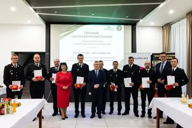 Grupa osób, głównie w mundurach strażackich, stoi podczas uroczystości wręczenia zaświadczeń w Sułkowicach, związanej z otwarciem zmodernizowanej kuchni w ramac