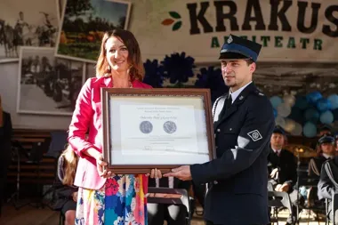 Kobieta w różowej marynarce i mężczyzna w mundurze trzymają wspólnie oprawiony dyplom na scenie z napisem „KRAKUS AMITEATR”.