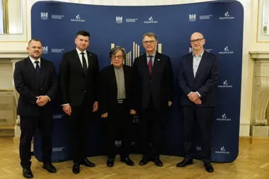 Pięć osób pozuje do grupowego zdjęcia przed niebieskim ściankiem z logo Instytucji Kultury Miasta Krakowa i Małopolski w eleganckim pomieszczeniu.