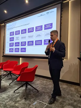 Mężczyzna w garniturze przemawia do mikrofonu podczas prezentacji slajdu o powstaniu Zintegrowanej Sieci Kolejowej w sali konferencyjnej z czerwonymi fotelami.