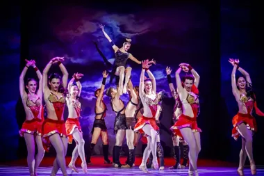 Zespół tancerzy w kostiumach scenicznych wykonuje choreografię na scenie, a jedna z tancerek unoszona jest w górę przez partnerów.
