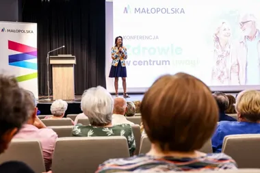 Kobieta przemawia z mikrofonem na scenie podczas konferencji Małopolski o zdrowiu, siedząca publiczność słucha prelegentki.