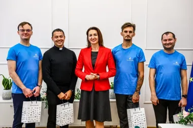 Grupa pięciu osób, w tym kobieta w czerwonej marynarce i mężczyzna w czarnym duchownym ubraniu, pozuje do zdjęcia w eleganckim pomieszczeniu; trzej mężczyźni w