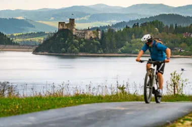Rowerzysta jedzie drogą nad jeziorem, w tle na wzgórzu widać średniowieczny zamek otoczony lasem i góry.