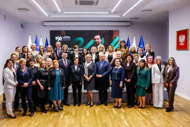 Grupa osób pozuje do pamiątkowego zdjęcia w sali konferencyjnej z flagami polskimi i unijnymi oraz portretem na ekranie w tle.