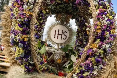 Dekoracja dożynkowa z kłosów zbóż, suszonych kwiatów i nasion z monogramem IHS oraz koszem, ustawiona na zewnątrz w słoneczny dzień.