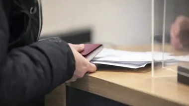 Dłonie osoby trzymającej paszport i dokumenty na drewnianym blacie okienka urzędowego.