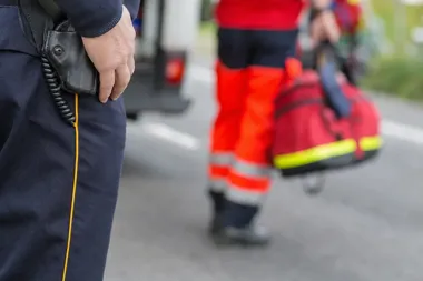 Funkcjonariusz w granatowym mundurze z radiotelefonem stoi na drodze, w tle ratownik w pomarańczowym ubiorze niesie czerwoną torbę medyczną.
