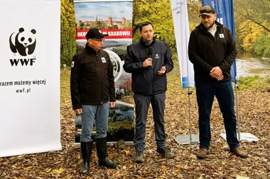 Trzech mężczyzn stoi podczas wydarzenia plenerowego nad brzegiem rzeki w jesiennym lesie, jeden z nich przemawia do mikrofonu obok banerów WWF i promujących Kra