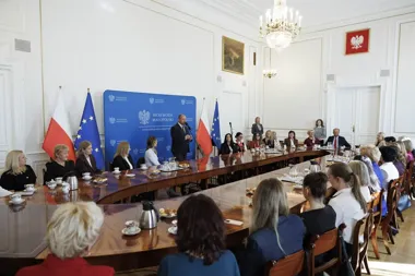 W sali urzędu wojewódzkiego w Małopolsce mężczyzna przemawia do grupy osób zasiadających przy dużym stole konferencyjnym, za nim stoją flagi Polski i UE oraz ba