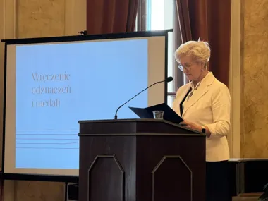 Kobieta w białym żakiecie stoi przy mównicy podczas uroczystości wręczenia odznaczeń i medali, w tle ekran z napisem "Wręczenie odznaczeń i medali".