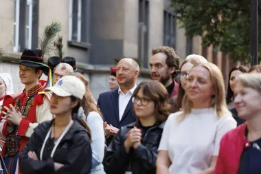Grupa ludzi, w tym osoba w stroju ludowym z pawimi piórami na kapeluszu, stoi na zewnątrz przed budynkiem i bije brawo.