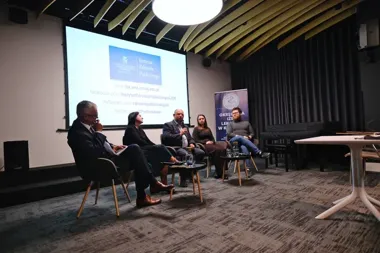 Panel dyskusyjny z pięcioma uczestnikami siedzącymi na krzesłach w sali konferencyjnej, z projekcją Instytutu Zdrowia Publicznego UJ CM na ekranie w tle.