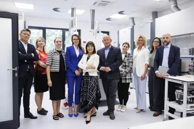 Grupa osób w eleganckim stroju biznesowym pozuje do zdjęcia w nowoczesnym laboratorium z okapami wyciągowymi i sprzętem laboratoryjnym.
