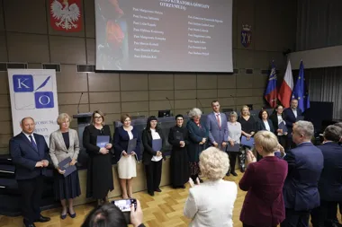 Uroczystość wręczenia nagród kuratora oświaty w sali konferencyjnej, podczas której wyróżnione osoby stoją na scenie z dyplomami, a publiczność im aplauduje.
